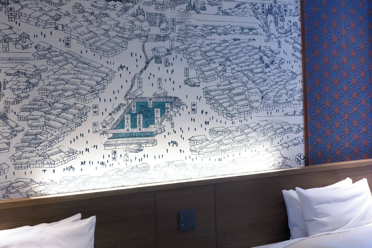 A hotel room with a large wall mural depicting a traditional town scene, a bed with white linens, and a patterned curtain.