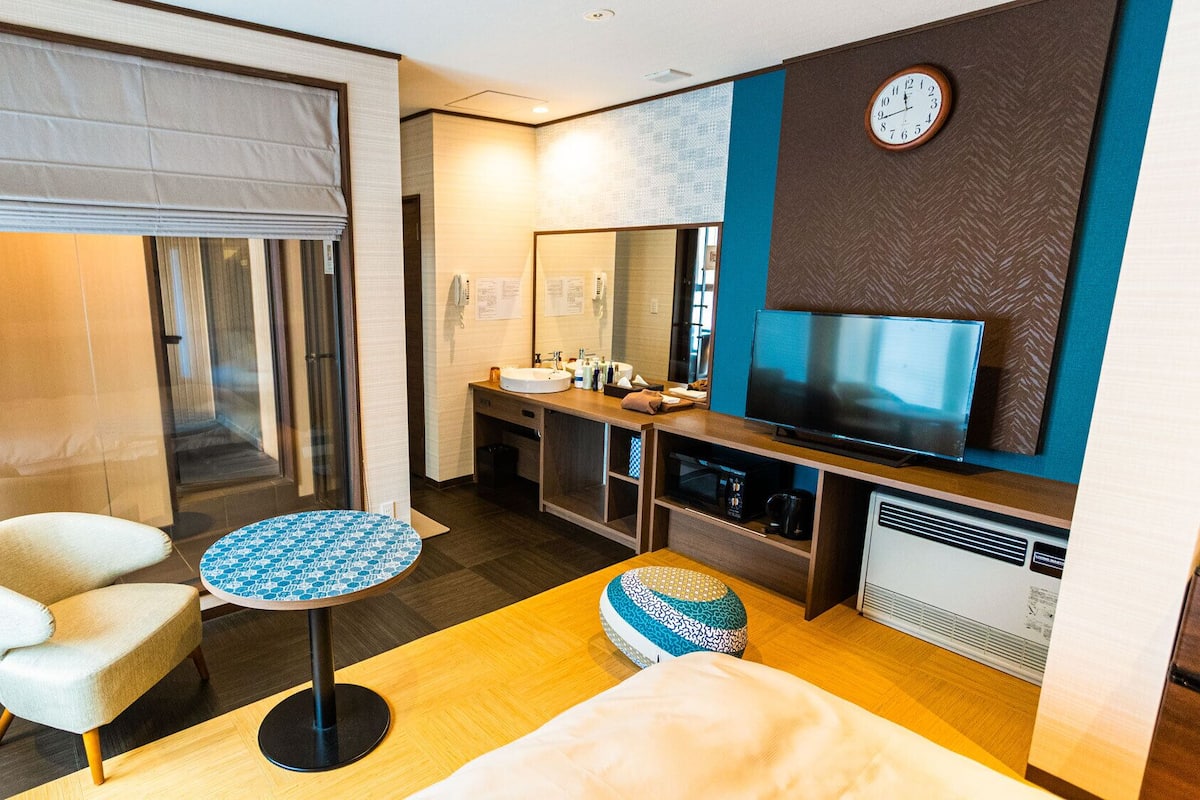 A hotel room with a bed, a round table, a TV, a bathroom with a sink and mirror, and a window with a view.