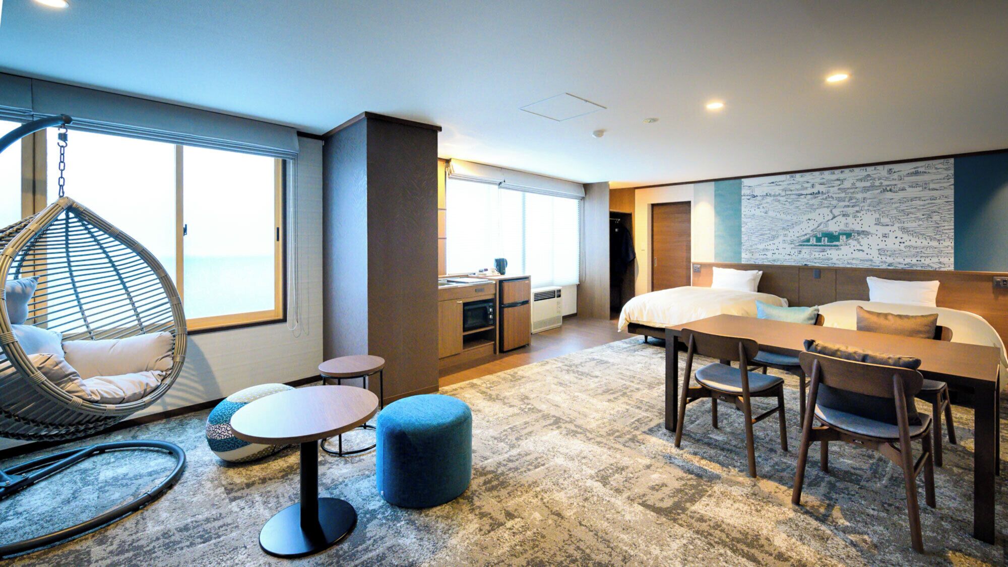 A modern hotel room with a bed, a dining table, a hanging chair, a small kitchen area, and a large window.