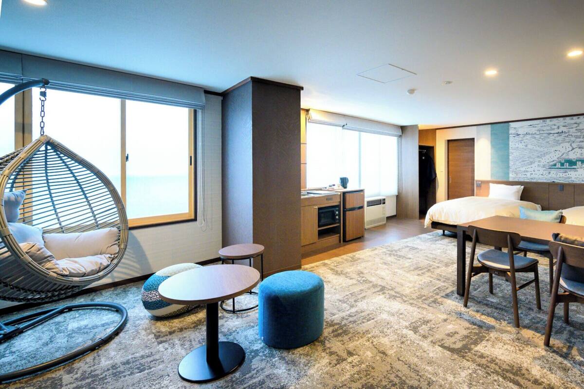 A modern hotel room with a bed, a dining table, a hanging chair, a small kitchen area, and a large window.