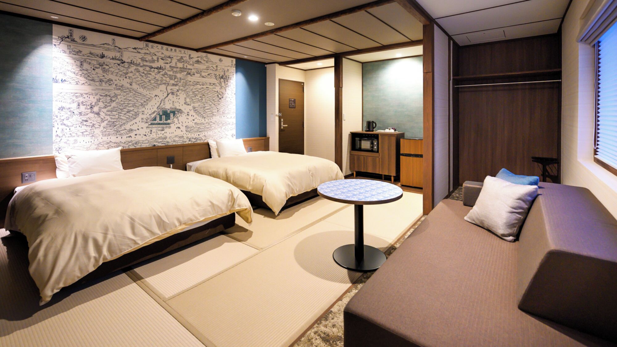 A hotel room with two beds, a small round table, a sofa, a map on the wall, and a kitchenette.