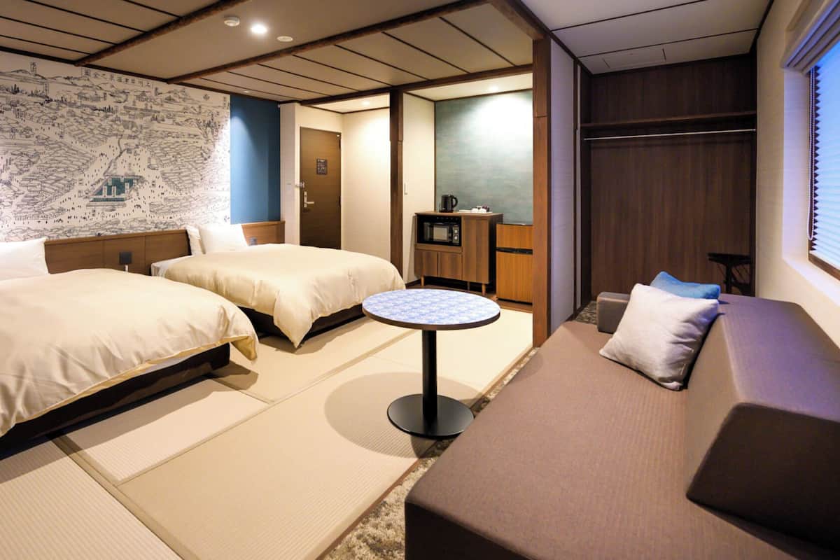 A hotel room with two beds, a small round table, a sofa, a map on the wall, and a kitchenette.