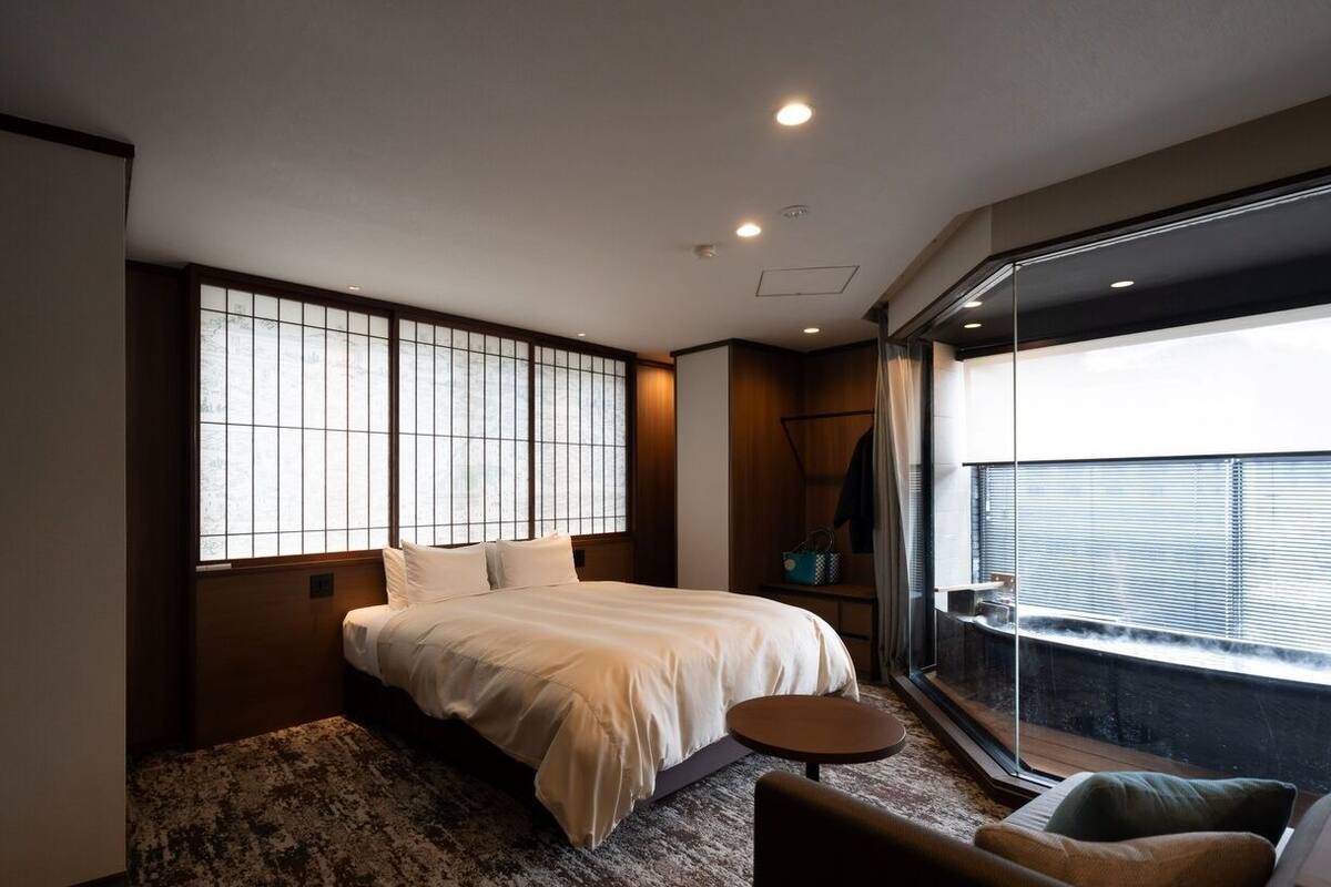 A modern hotel room with a large bed, a small round table, a sofa, and a view of the city through large windows.