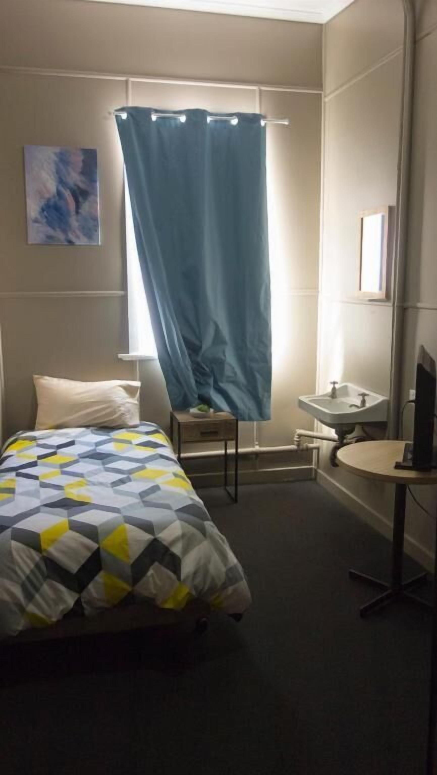 A hotel room with a bed, a small bedside table, a desk with a chair, a window with a curtain, and a bathroom with a sink and mirror.