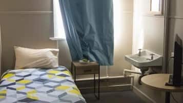 A hotel room with a bed, a small bedside table, a desk with a chair, a window with a curtain, and a bathroom with a sink and mirror.