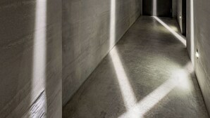 Un couloir étroit aux murs en béton, où une série de luminaires projette des ombres sur le sol.