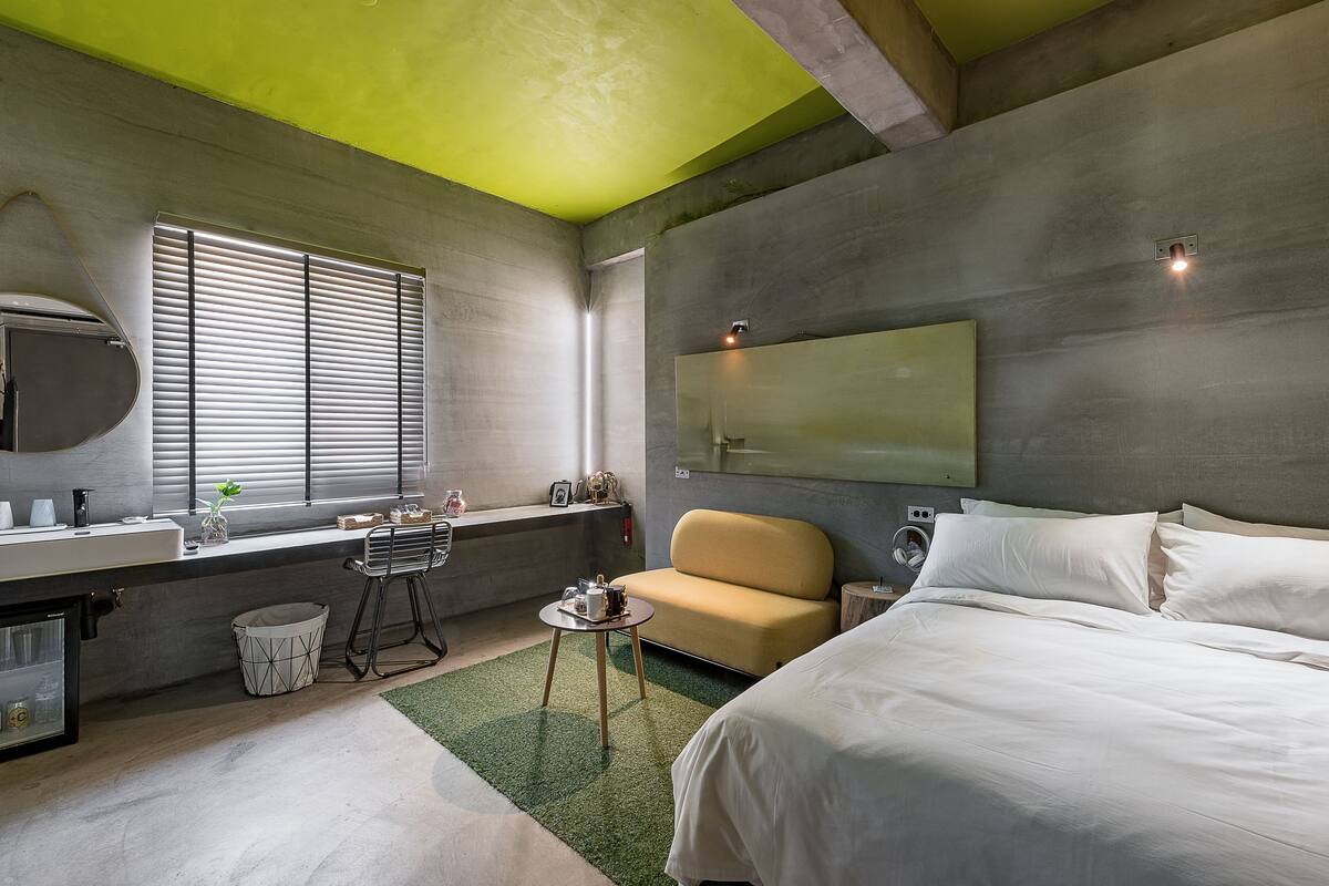 A modern hotel room with a bed, a sofa, a small table, a window with blinds, a mirror, and a green ceiling.
