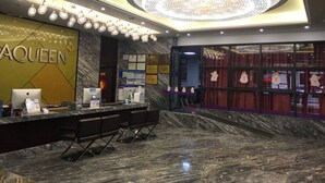 Un hall d’hôtel avec un sol en marbre, un grand lustre et un comptoir équipé d’un ordinateur et d’une enseigne sur laquelle on peut lire « AQUEEN ».