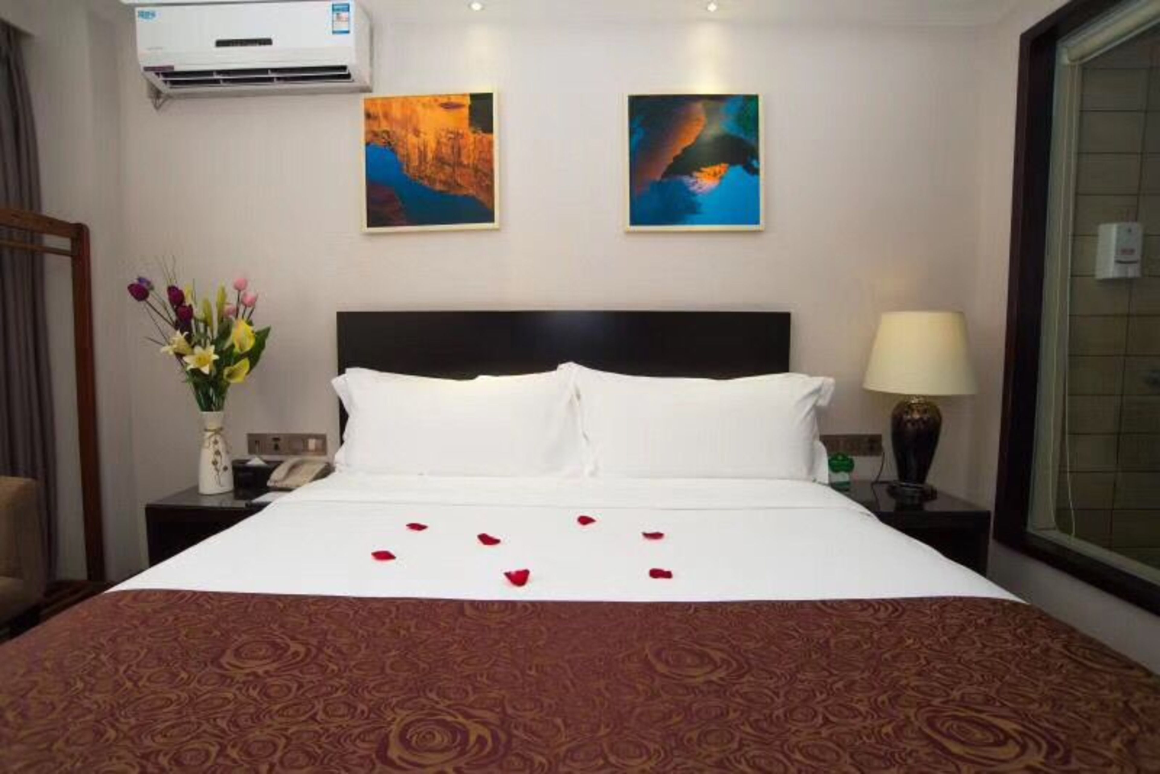 A hotel room with a large bed, two paintings on the wall, a vase of flowers, and a nightstand with a lamp and a phone.
