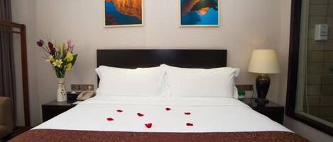 A hotel room with a large bed, two paintings on the wall, a vase of flowers, and a nightstand with a lamp and a phone.