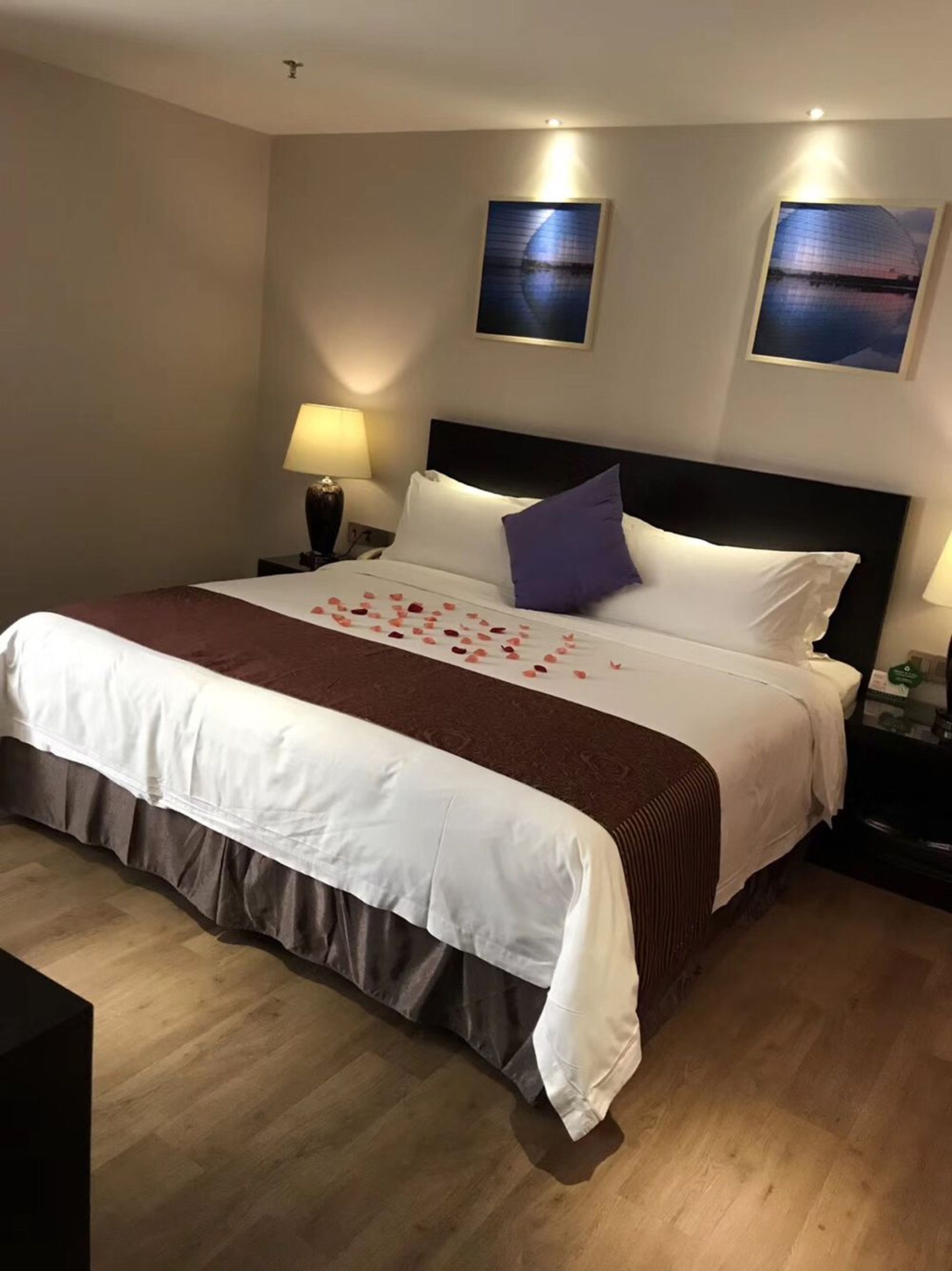 A neatly made bed with a white and brown bedspread, a purple pillow, and rose petals on the bed. There are two wall-mounted lamps and two framed pictures on the wall.