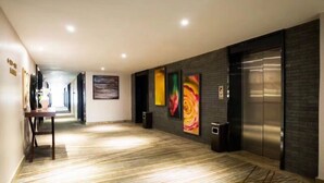 Un couloir avec un tapis à motifs, une table sur laquelle est posé un vase et un haut-parleur. Il y a quatre tableaux colorés accrochés au mur et un grand ascenseur.