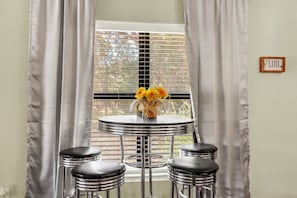 A room with a round table, two stools, a vase with flowers, and a window with blinds.
