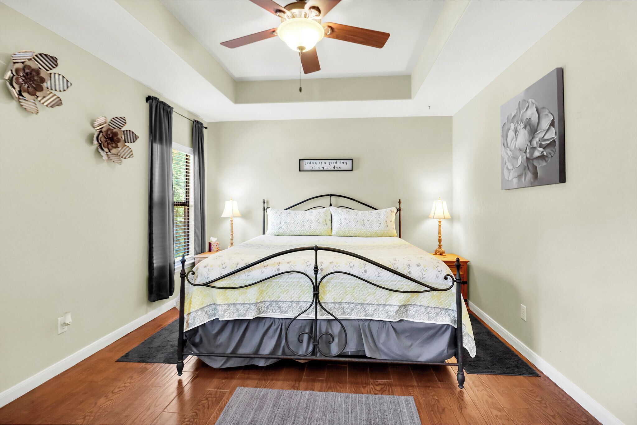 A bedroom with a bed, two bedside lamps, a ceiling fan, a window with curtains, a wall clock, and a framed quote on the wall.