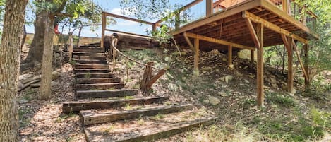 Una escalera de madera conduce a una plataforma también de madera, con cerramientos de vidrio, rodeada de árboles y vegetación natural.