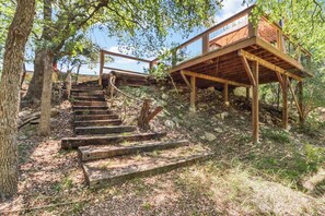 Una escalera de madera conduce a una plataforma también de madera, con cerramientos de vidrio, rodeada de árboles y vegetación natural.