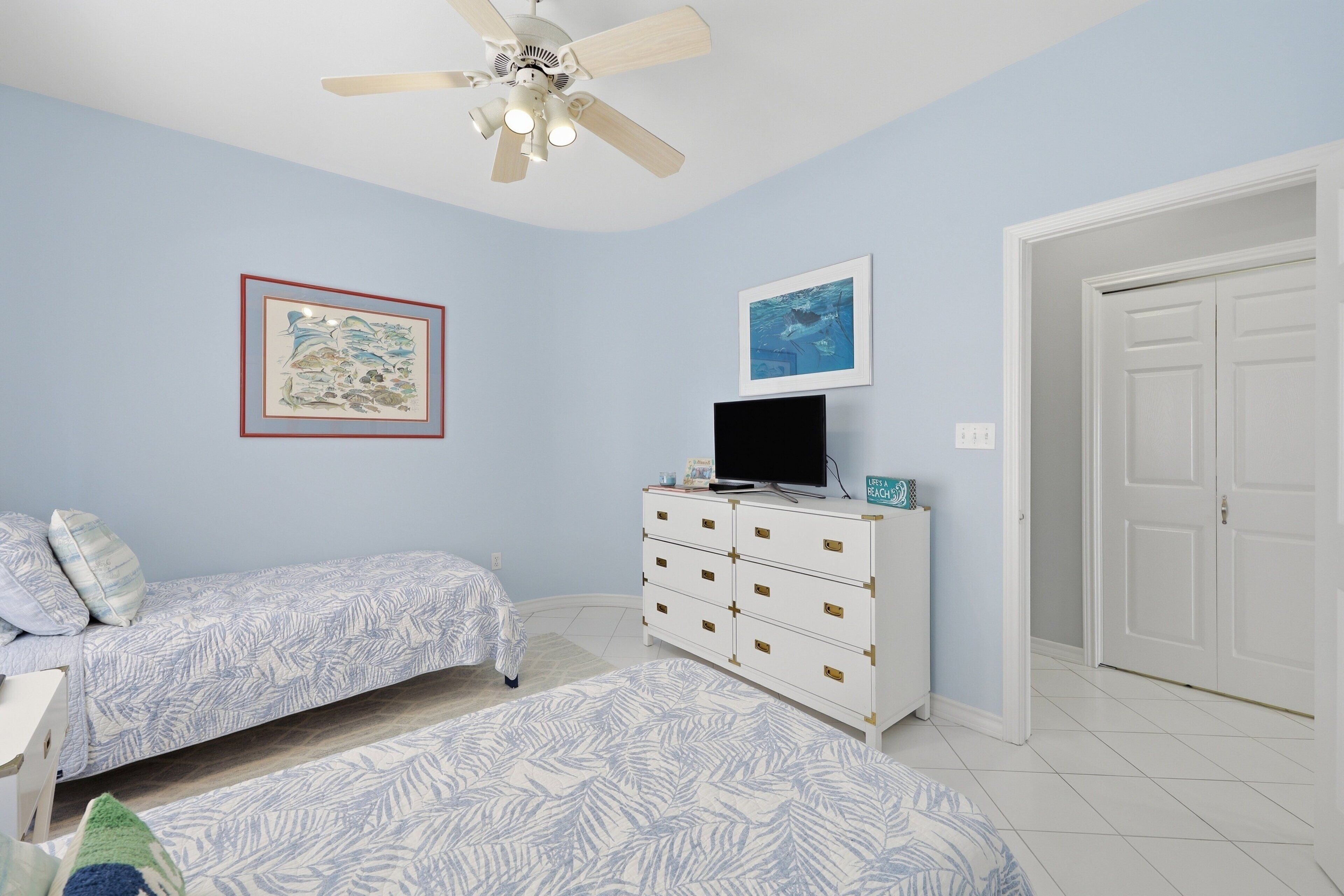 A bedroom with two beds, a dresser, a ceiling fan, and two framed pictures on the wall.