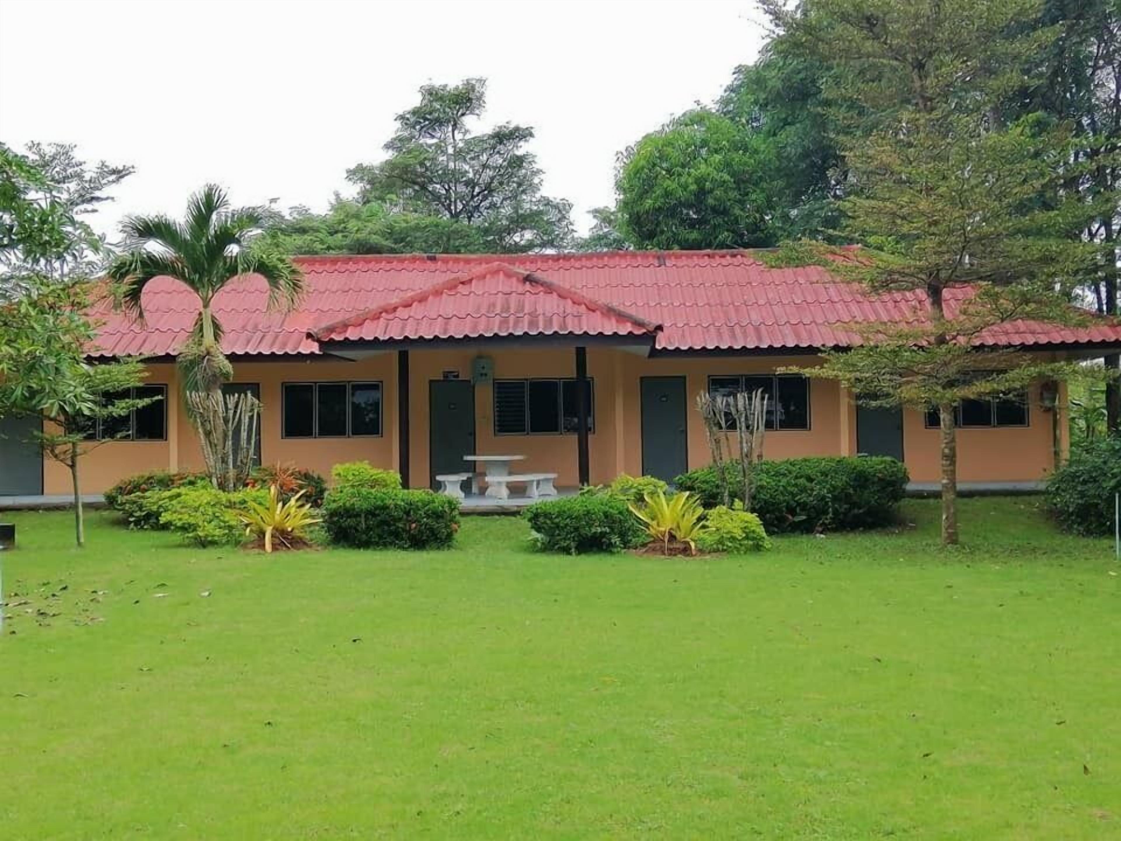 A two-story building with a red roof and beige walls, surrounded by a well-maintained lawn and various plants.