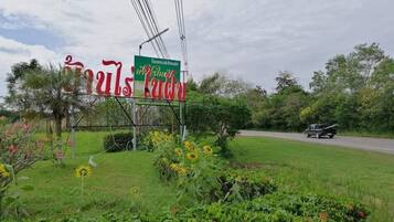 A roadside sign with Thai text, surrounded by greenery and a road with a vehicle.
