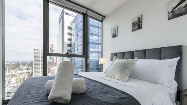 A modern hotel room with a large bed, a view of the cityscape, and a city skyline visible through the window. - Trendy Melbourne 2 Bedroom Apartment by BnB Pro (Melbourne)