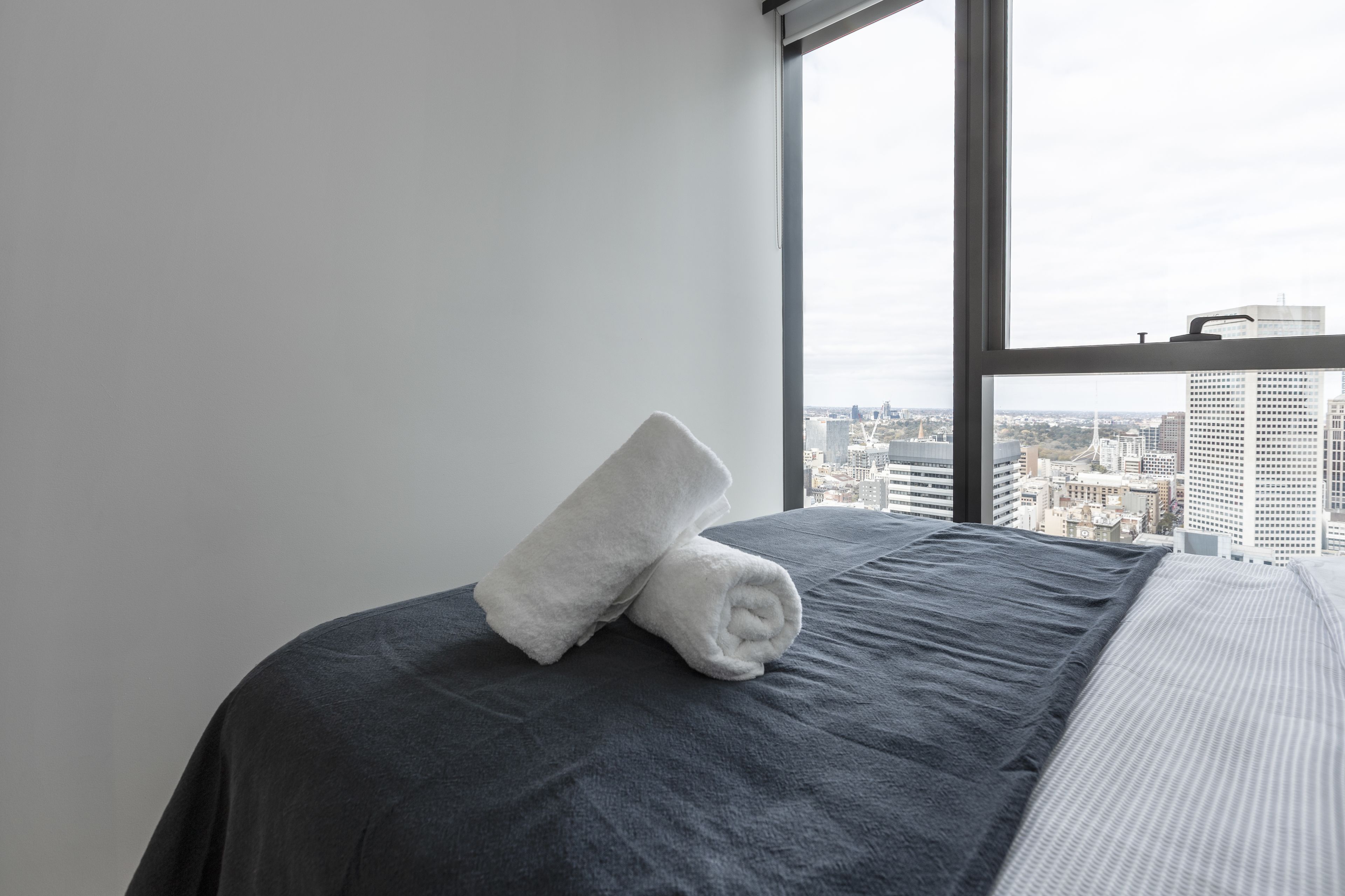 A neatly made bed with a dark gray bedspread and a white towel folded on top. A large window provides a view of a cityscape with tall buildings.
