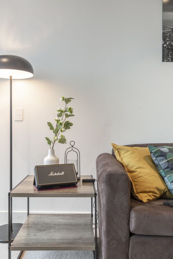 A modern living room with a sofa, a small side table, a lamp, a vase with flowers, and a Marshall speaker.