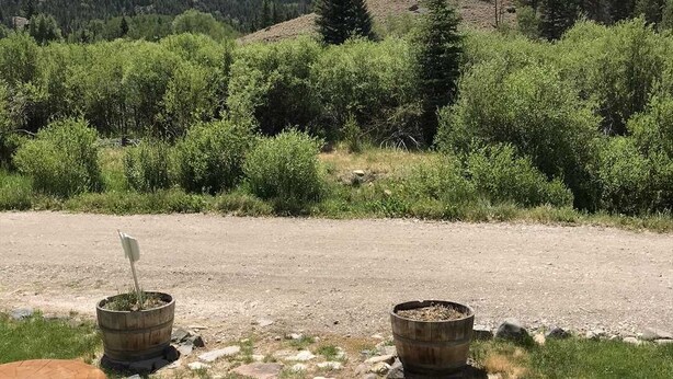 A wooden deck overlooking a gravel path with two rustic planters, surrounded by lush greenery and a mountain backdrop. - Redcloud Getaway - Great Property Along the Lake Fork of the Gunnison with Direct Access to Trails (Lake City)