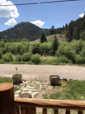 Una terraza de madera con vista a un sendero de grava y dos macetas rústicas, rodeada de abundante vegetación y un telón de fondo montañoso.