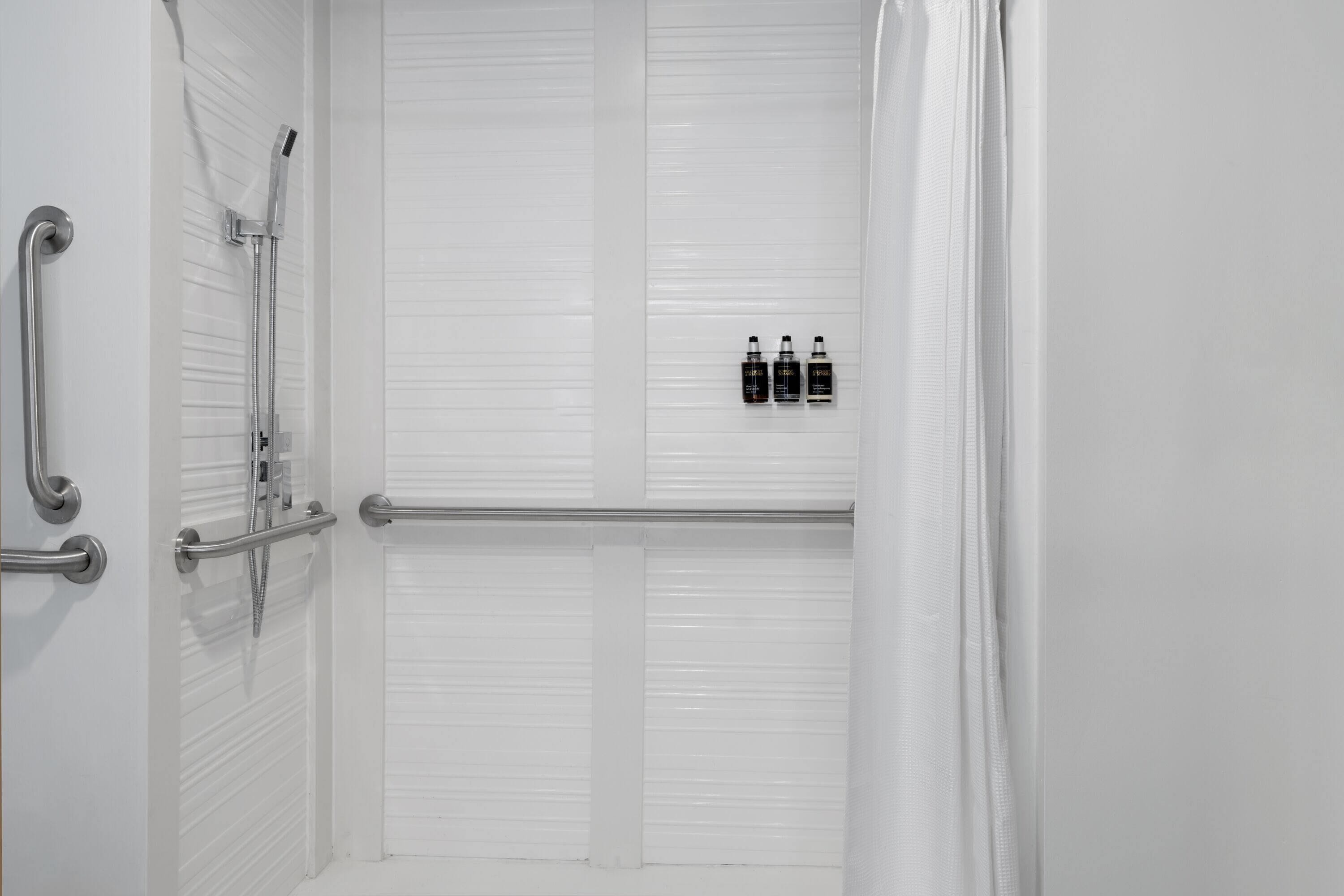 A white bathroom with a shower, grab bars, and three bottles of shampoo on the wall.
