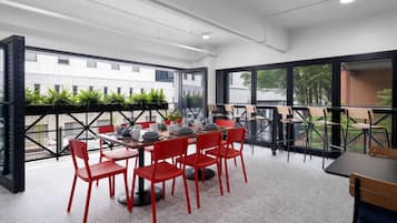 A modern dining area with red chairs and a wooden table, large windows with black frames, and a view of buildings and greenery outside.