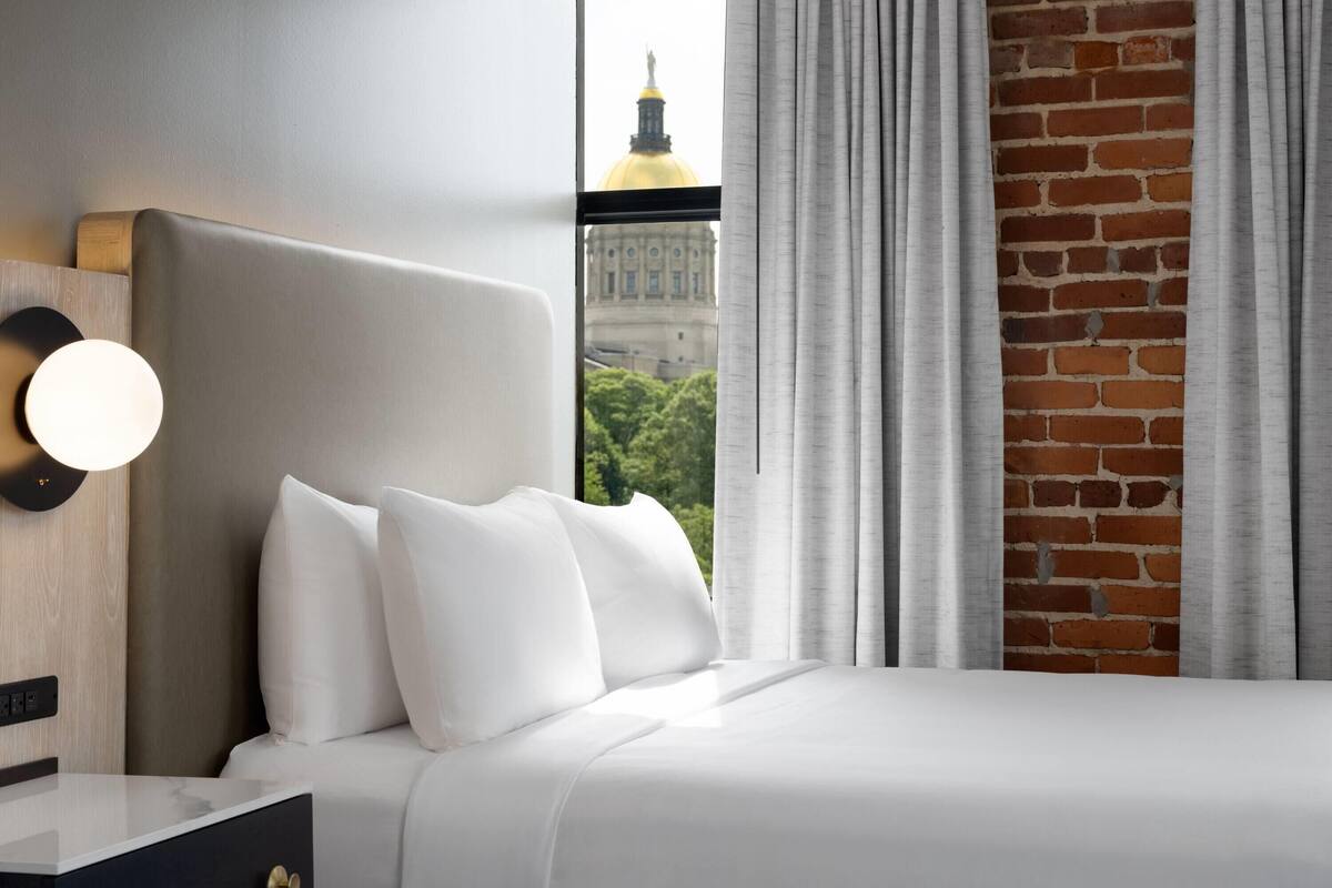 A hotel room with a large bed, a nightstand, a wall-mounted light, and a view of a building through a window with curtains.