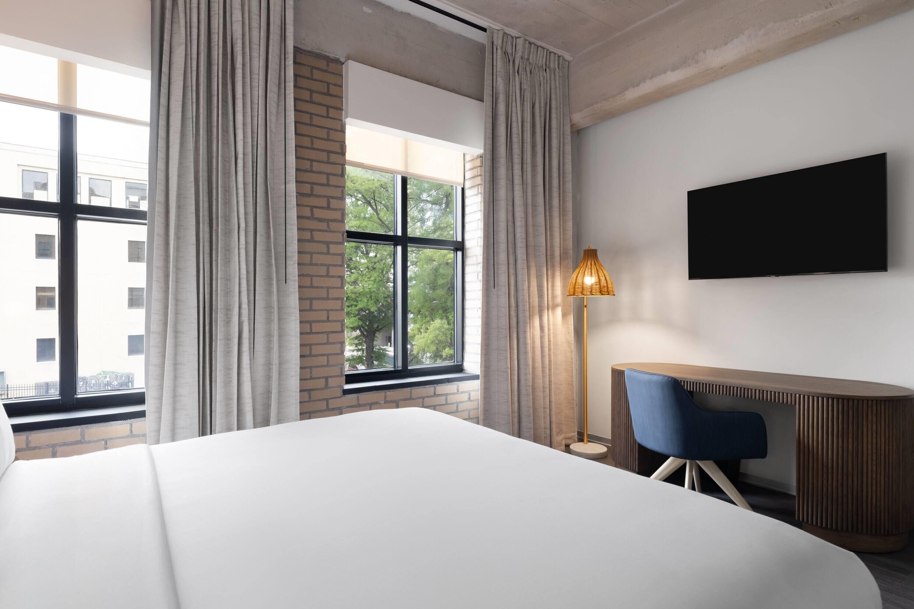 Une chambre d’hôtel avec un grand lit, un bureau avec une chaise, un téléviseur fixé au mur et une fenêtre avec des rideaux.