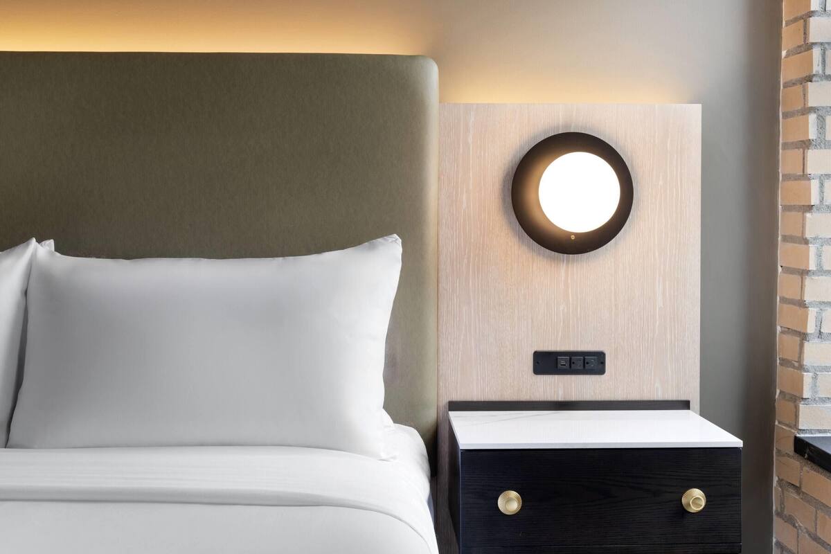 A neatly made bed with white linens, a round wall-mounted light, and a small wooden nightstand with a digital clock.