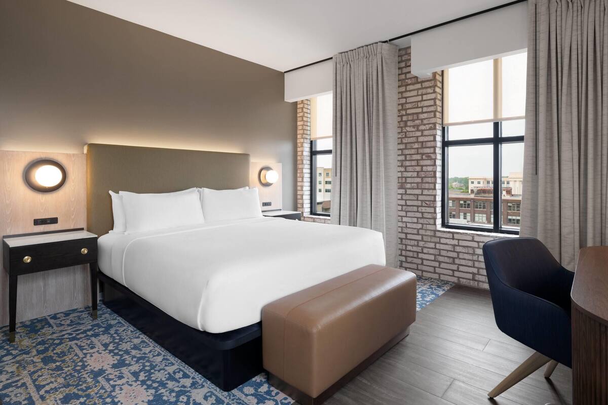 A hotel room with a large bed, a nightstand, a bench, a chair, and a view of buildings through a window.