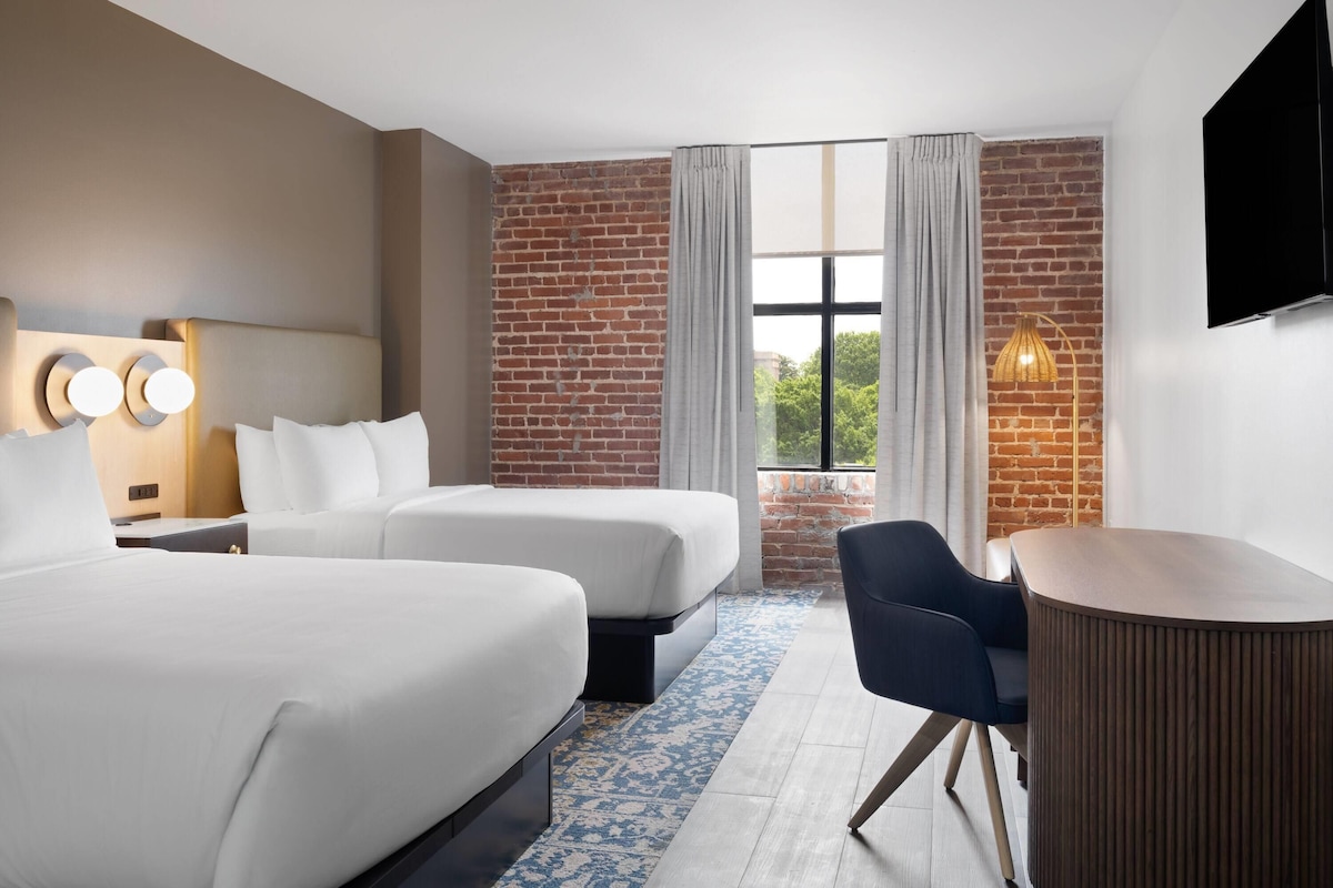 A hotel room with two beds, a desk, a chair, a TV, and a large window with curtains.
