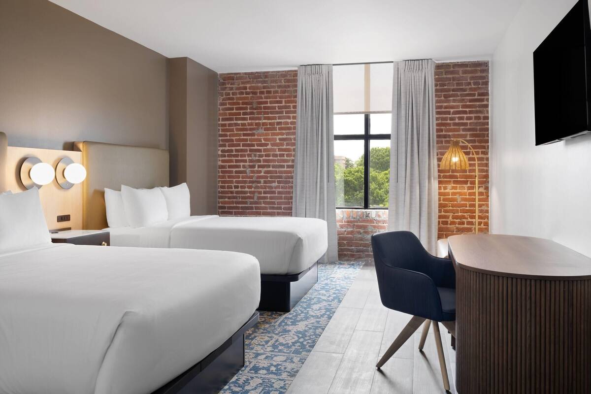 A hotel room with two beds, a desk, a chair, a TV, and a window with curtains.
