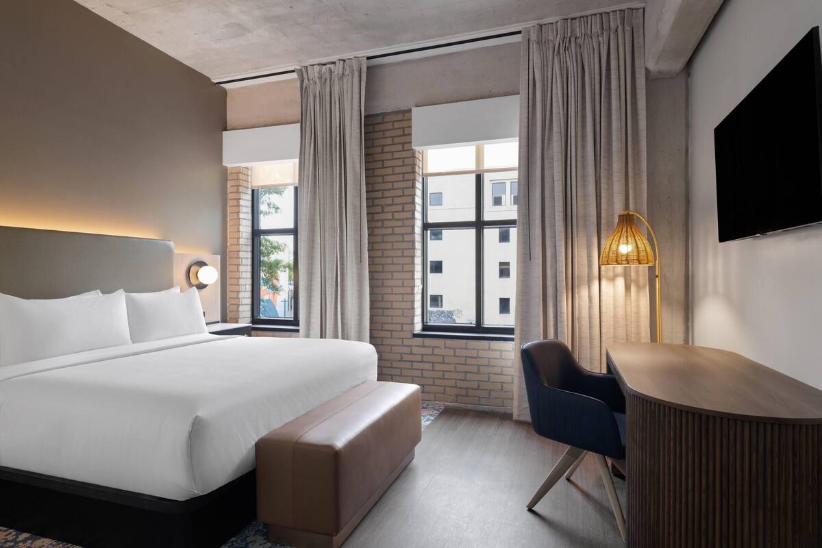 A modern hotel room with a large bed, a desk, a chair, and a TV on the wall.