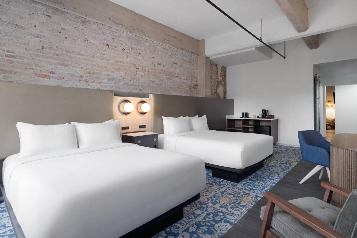 A hotel room with two beds, a desk, a chair, a small kitchenette, and a brick wall.