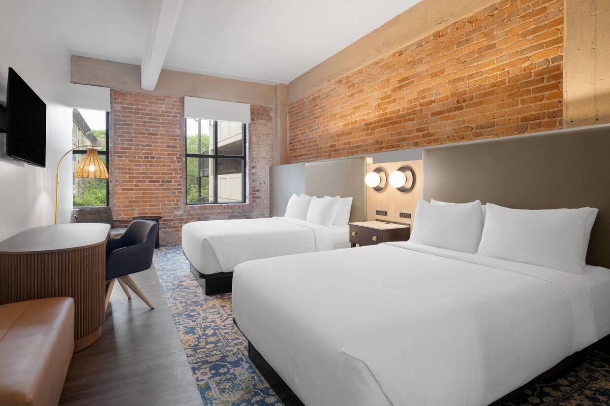 A hotel room with two beds, a TV, a brick wall, and a large window.