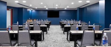 A conference room with rows of tables and chairs, a large screen, and water bottles on each table.