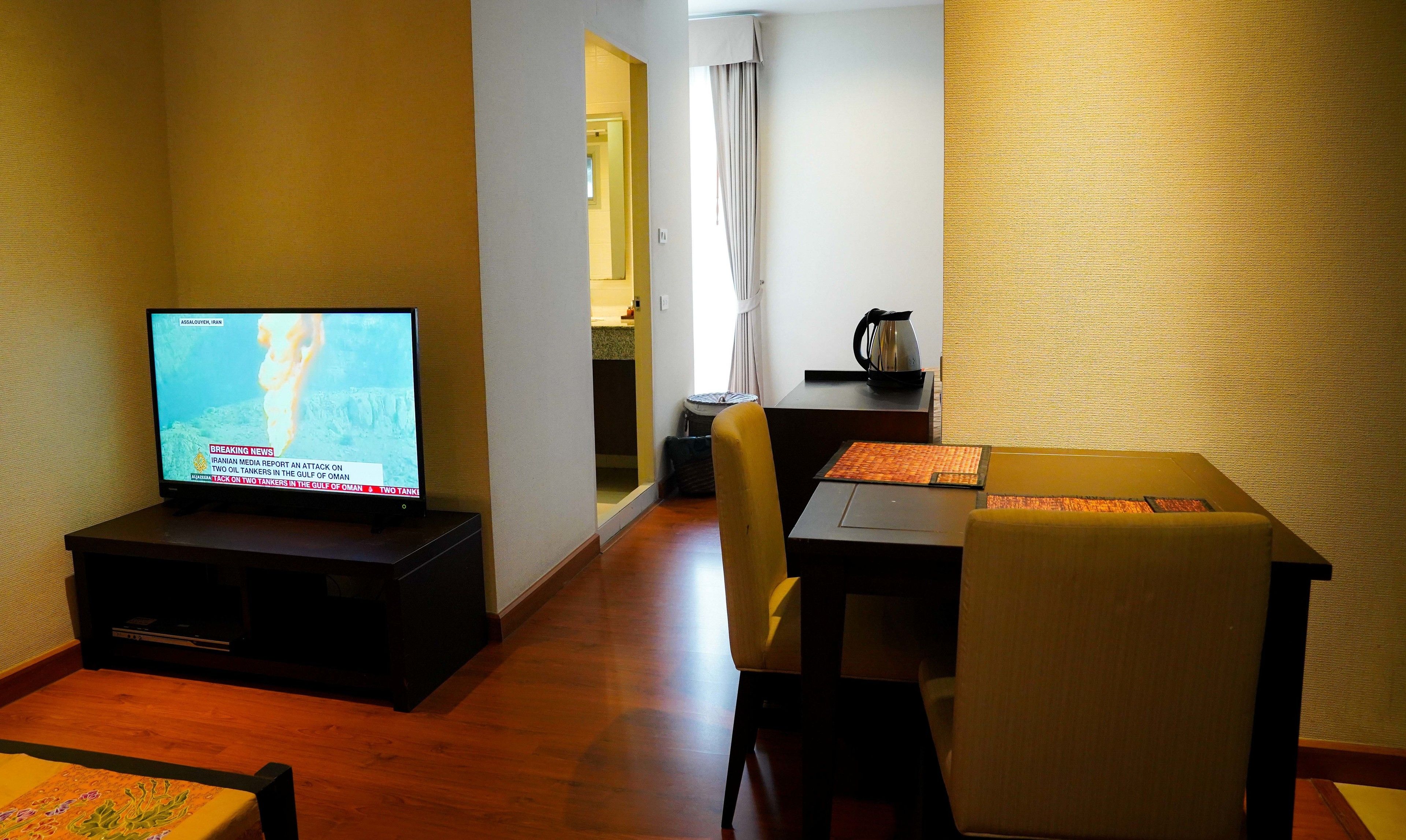 Habitación de hotel con televisor mostrando noticias, zona de comedor con mesa y sillas, y cocina visible a través de una puerta abierta.