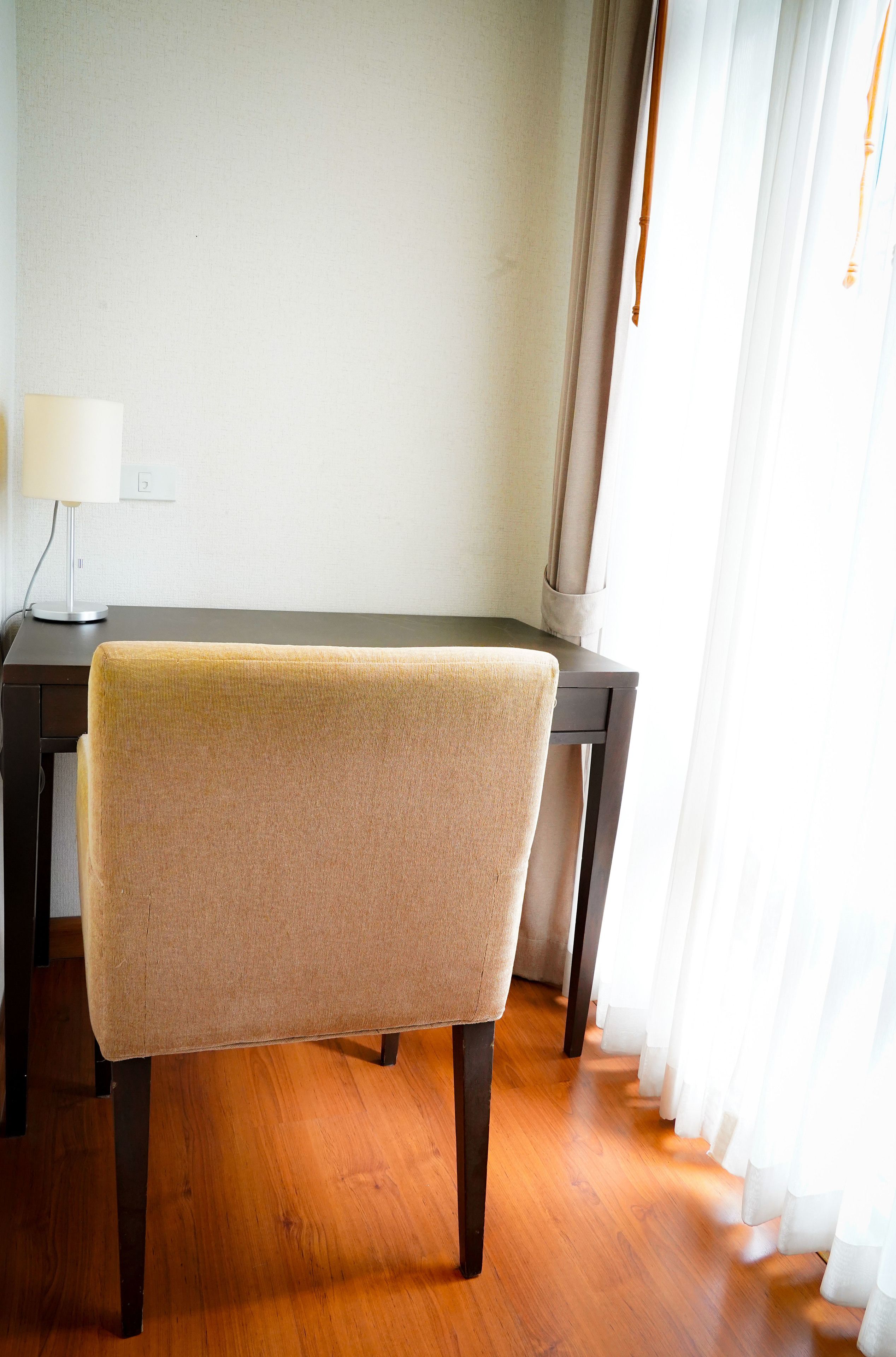 A chair with a beige cushion in a room with wooden flooring, a desk, and a lamp.