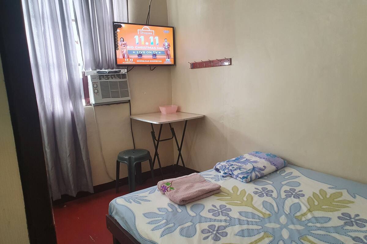 A small hotel room with a bed, a desk, a chair, a TV displaying a Christmas sale, and an air conditioner.