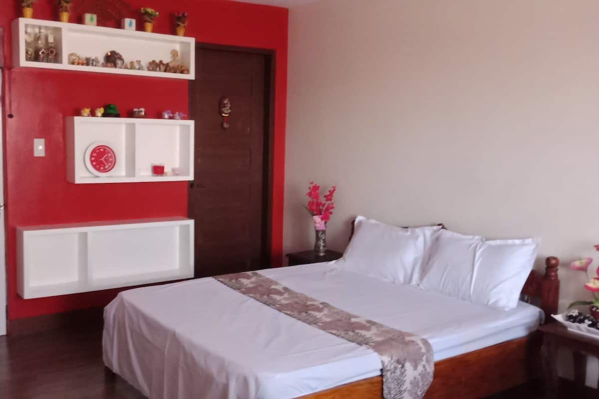 A bedroom with a bed, a white bedside table, a nightstand, a shelf with decorative items, and a door with a curtain.