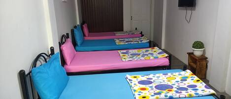 A corridor with four single beds, each with a different colored mattress and pillow. There is a potted plant on a wooden stool to the right.