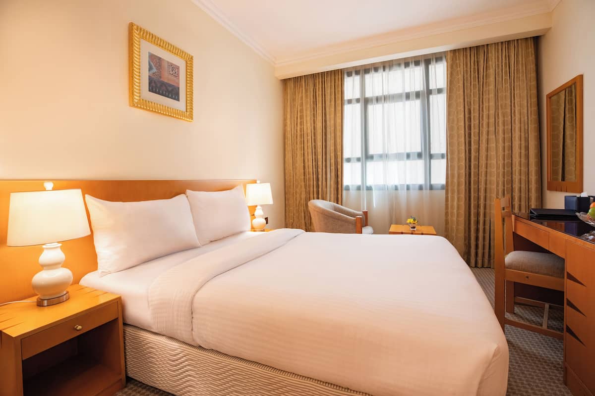 A hotel room with a large bed, two bedside tables with lamps, a desk with a chair, a large window with curtains, and a framed picture on the wall.