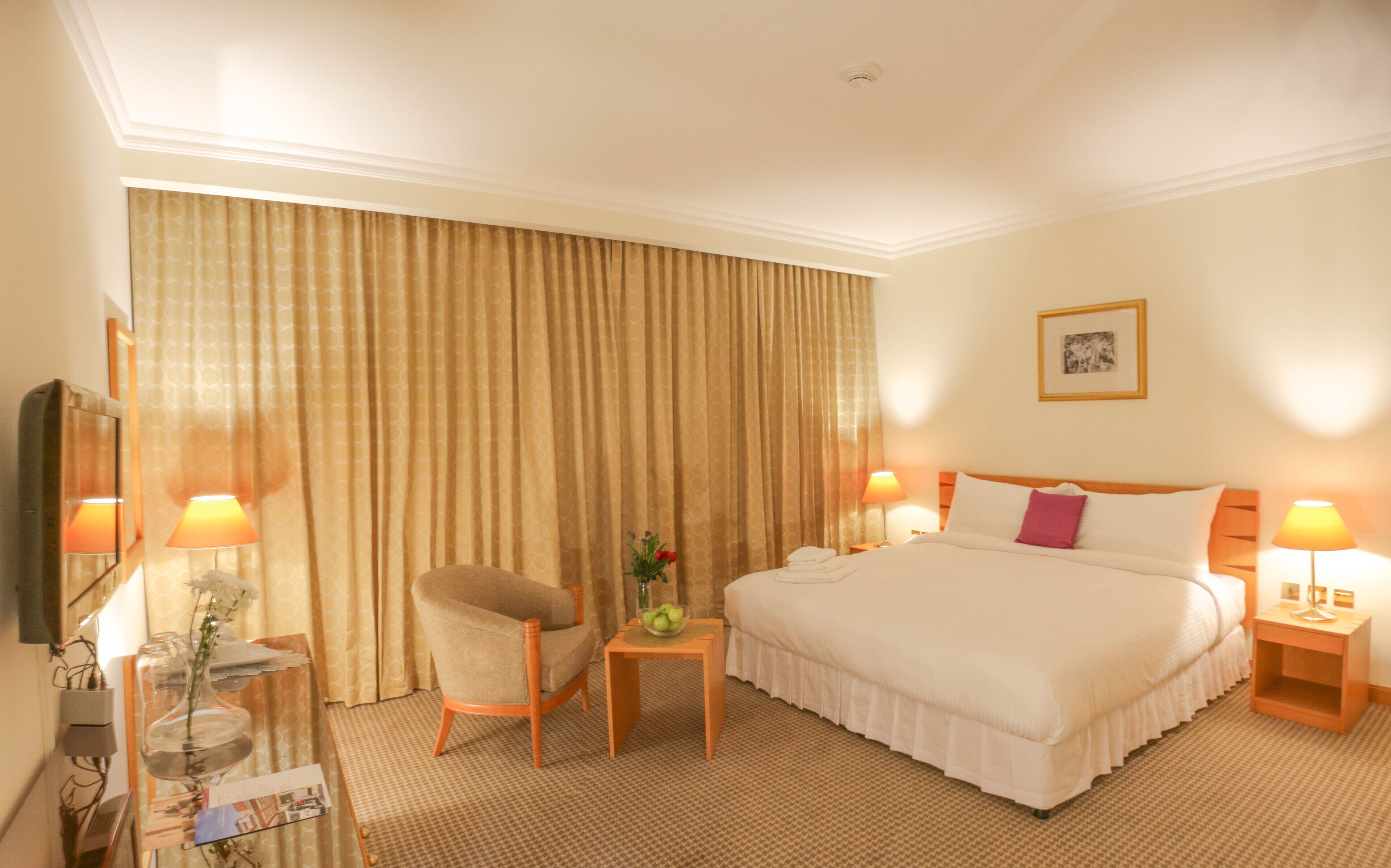 Ein Hotelzimmer mit einem großen Bett, zwei Nachttischen mit Lampen, einem Sessel, einem kleinen Tisch mit Blumenvase und einem großen Fenster mit Vorhängen.