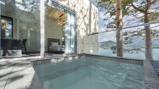 A modern outdoor pool area with clear glass walls, a bed visible inside, and a view of a lake and mountains. - Goseong Hansan Marina Resort (Goseong)