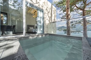 A modern outdoor pool area with clear glass walls, a bed visible inside, and a view of a lake and mountains. - Goseong Hansan Marina Resort (Goseong)