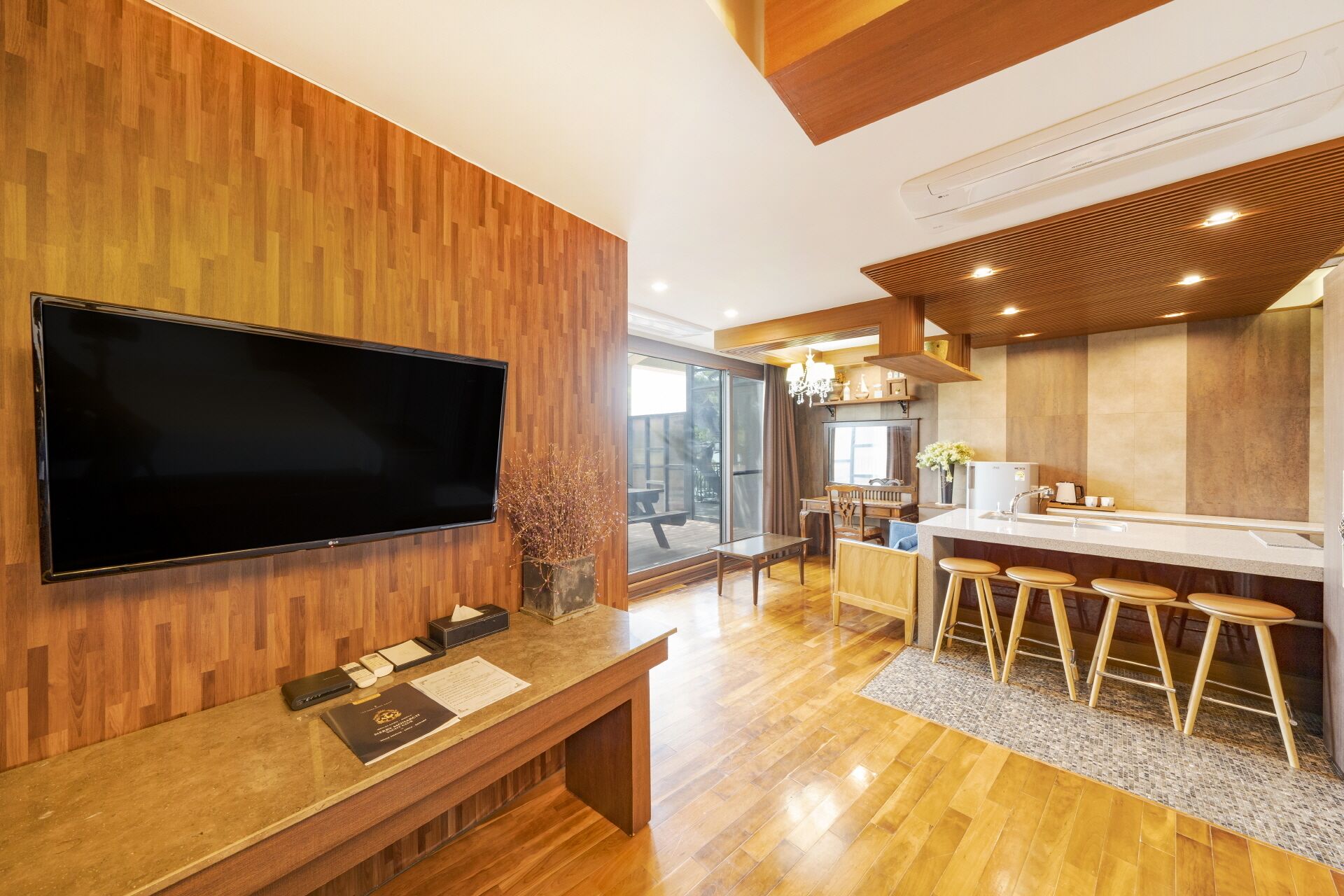 Una cocina moderna con piso de madera, un televisor de pantalla plana grande montado en una pared de madera, una barra con taburetes y una puerta corrediza de vidrio que da a un patio exterior.